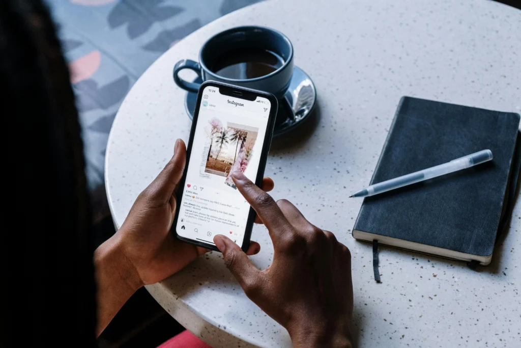 Uma pessoa segura um smartphone com a mão esquerda e usa o dedo indicador da mão direita para tocar na tela. O celular mostra uma postagem do Instagram com uma imagem de palmeiras e flores. Sobre a mesa branca e redonda, há uma xícara com café preto em um pires azul escuro e, ao lado, um caderno preto fechado com uma caneta transparente sobre ele.